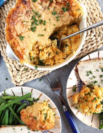 Overhead shot of cheesy corn casserole in a round dish with a silver spoon and two plates with turkey and green beans.