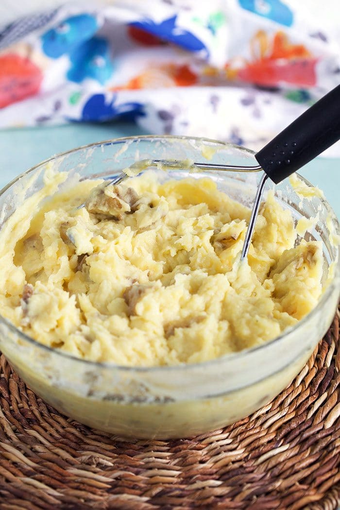 Mashed potatoes in a glass bowl with a potato masher.