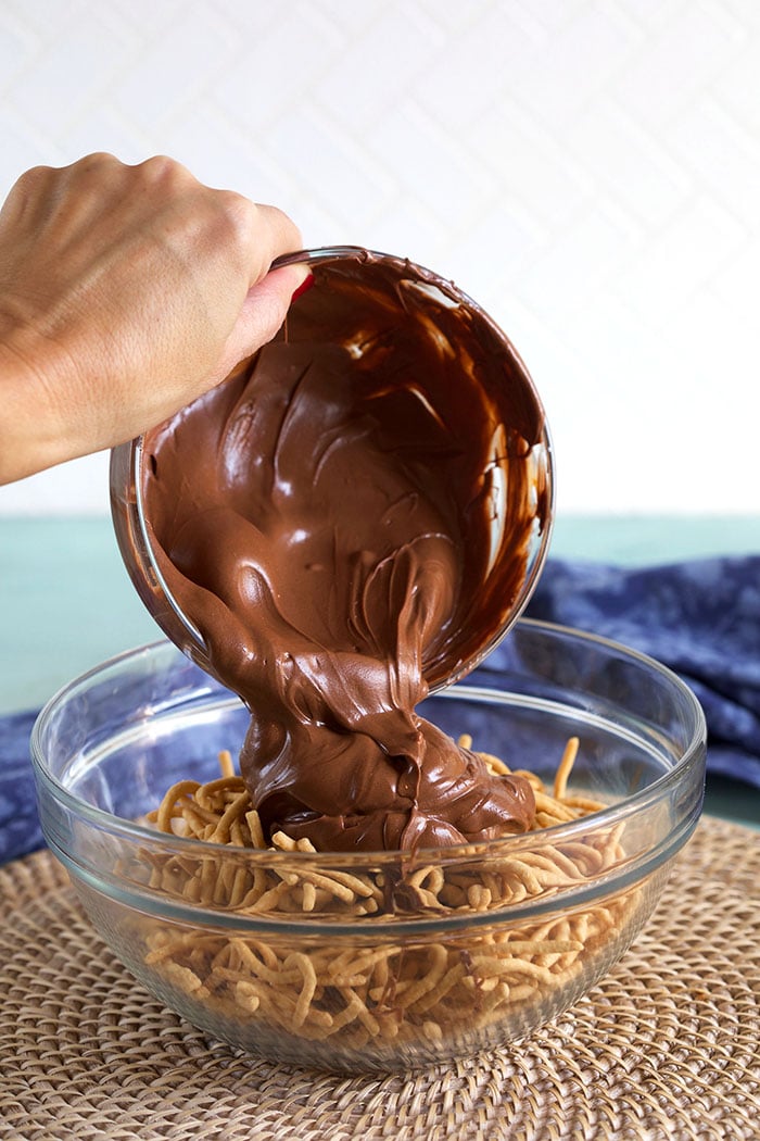Melted chocolate being poured over chow mein noodles in a glass bowl.