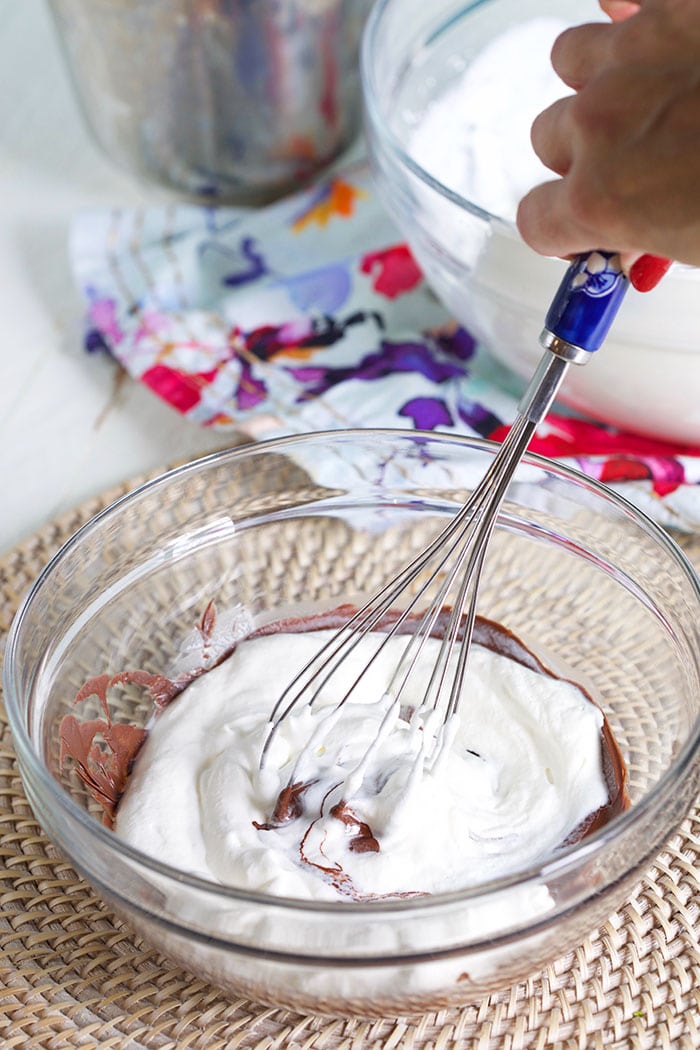 Whipped cream being whisked into melted chocolate in a glass bowl.
