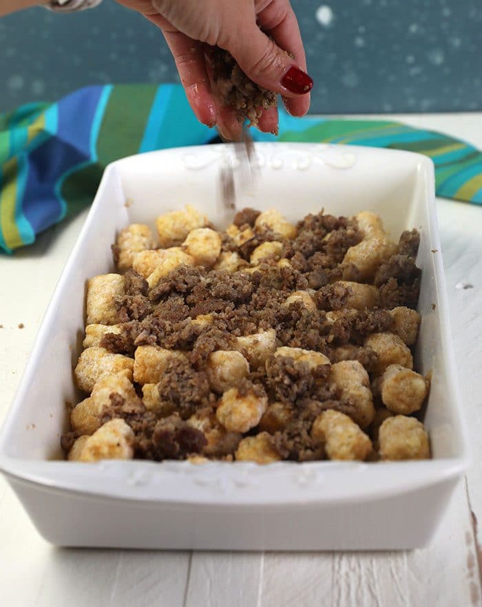 Tater tots in a casserole dish with sausage being sprinkled over it.