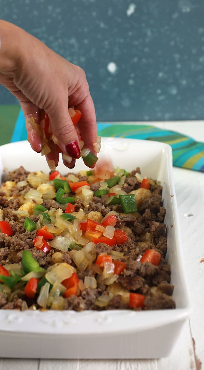 peppers and onions being sprinkled over tater tot casserole in a white baking dish.