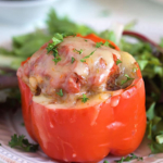 Stuffed red bell pepper topped with cheese on a white plate with greens.