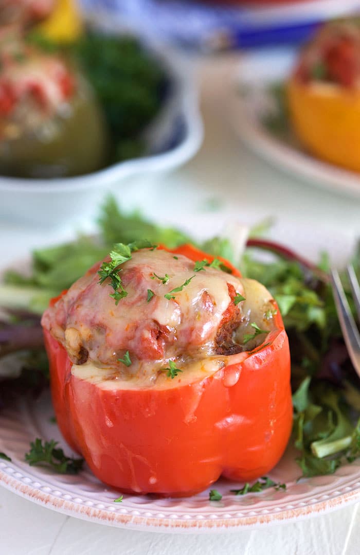 Stuffed red bell pepper topped with cheese on a white plate with greens.