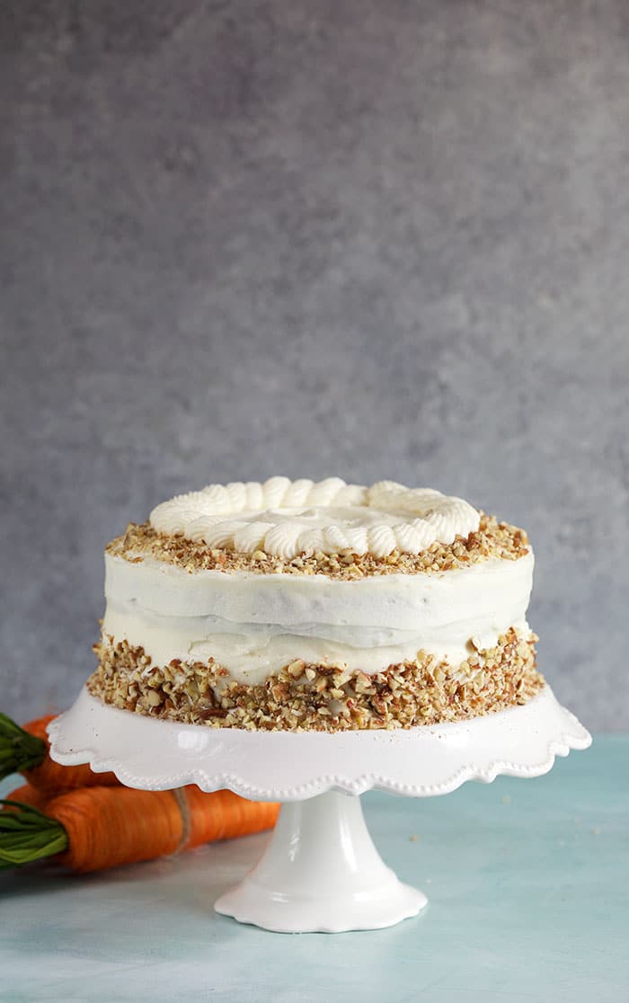 Whole carrot cake on a white cake pedestal on a gray background.