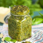 Basil Pesto in a glass jar with a wooden spoonful of basil pesto on a white striped napkin and basil leaves in the background