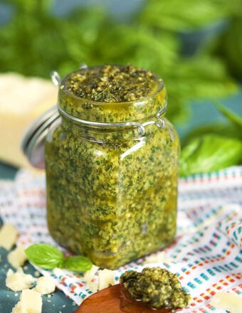Basil Pesto in a glass jar with a wooden spoonful of basil pesto on a white striped napkin and basil leaves in the background