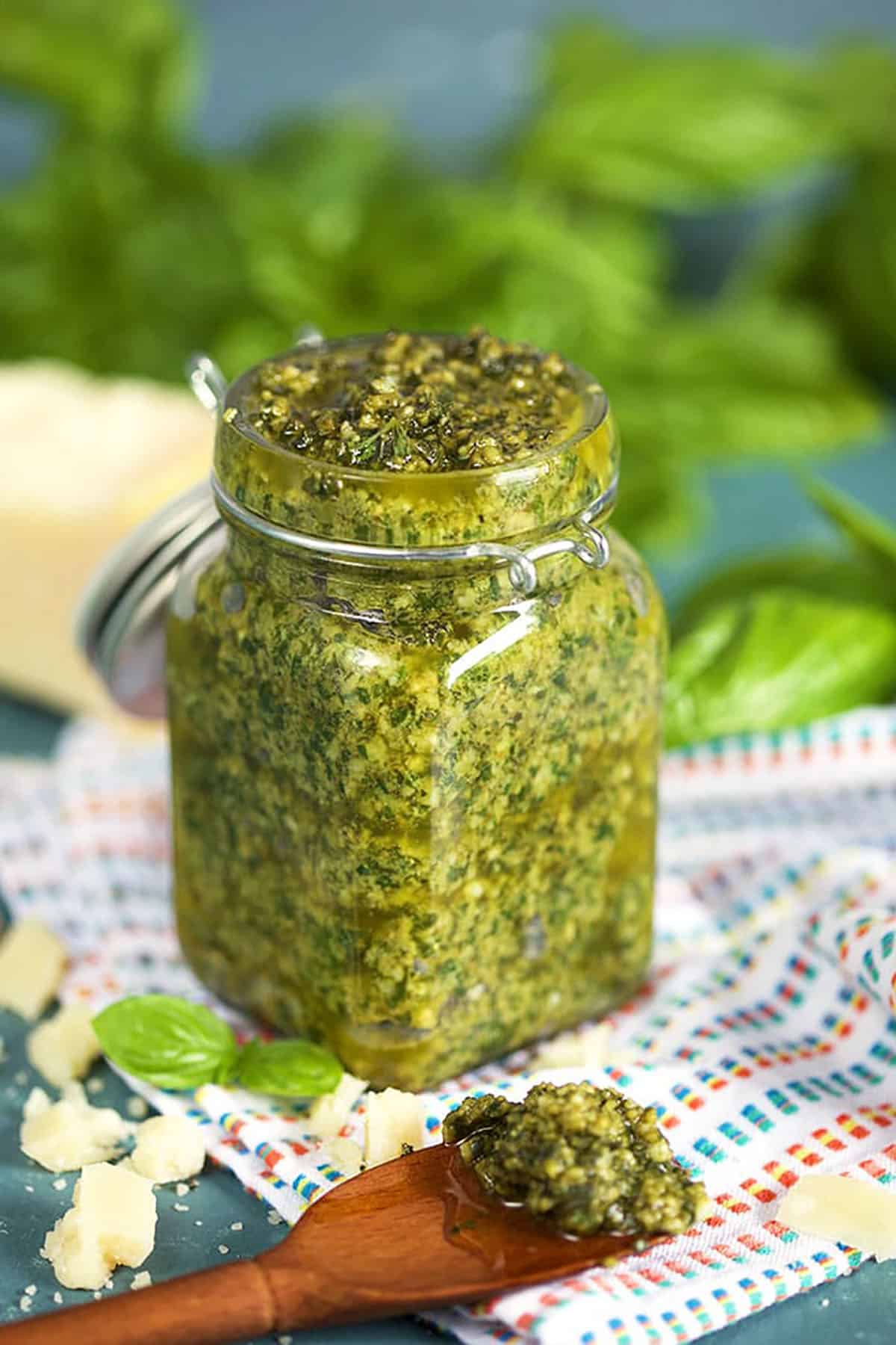 Basil Pesto in a glass jar with a wooden spoonful of basil pesto on a white striped napkin and basil leaves in the background 