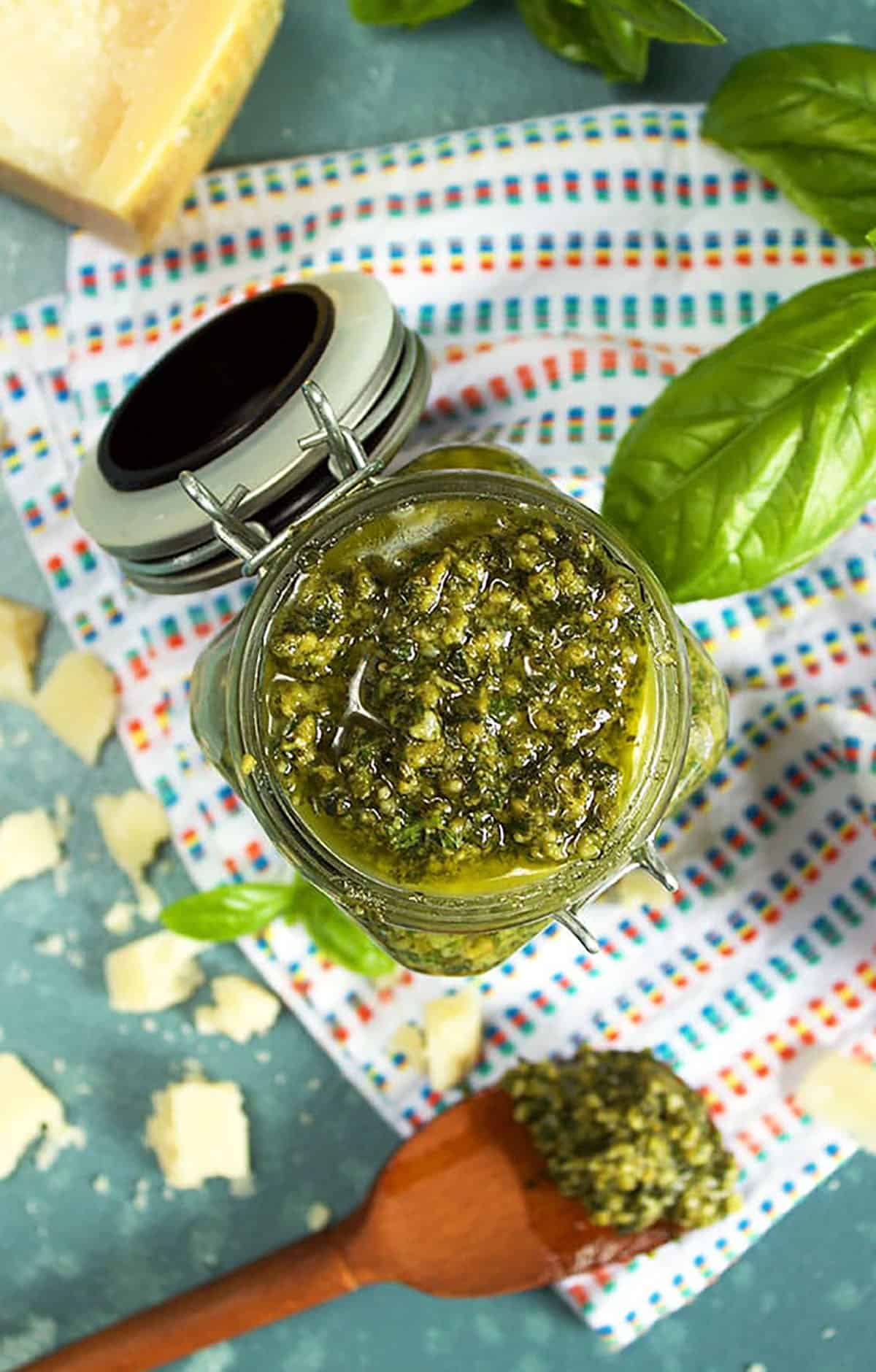 Overhead shot of the Very Best Basil Pesto in a glass jar with a teak spoon on a white napkin 