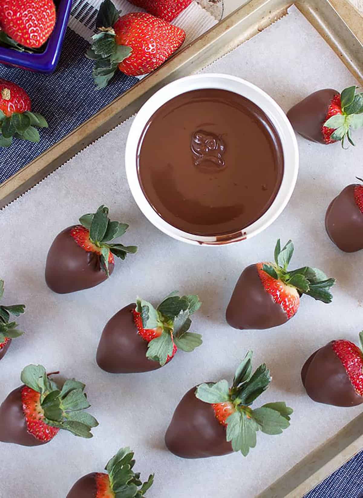 Bowl of melted chocolate on a baking sheet with strawberries dipped in chocolate