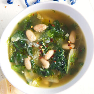 overhead shot of escarole soup with white beans in a white bowl with a blue tasseled towel near the top of the bowl.