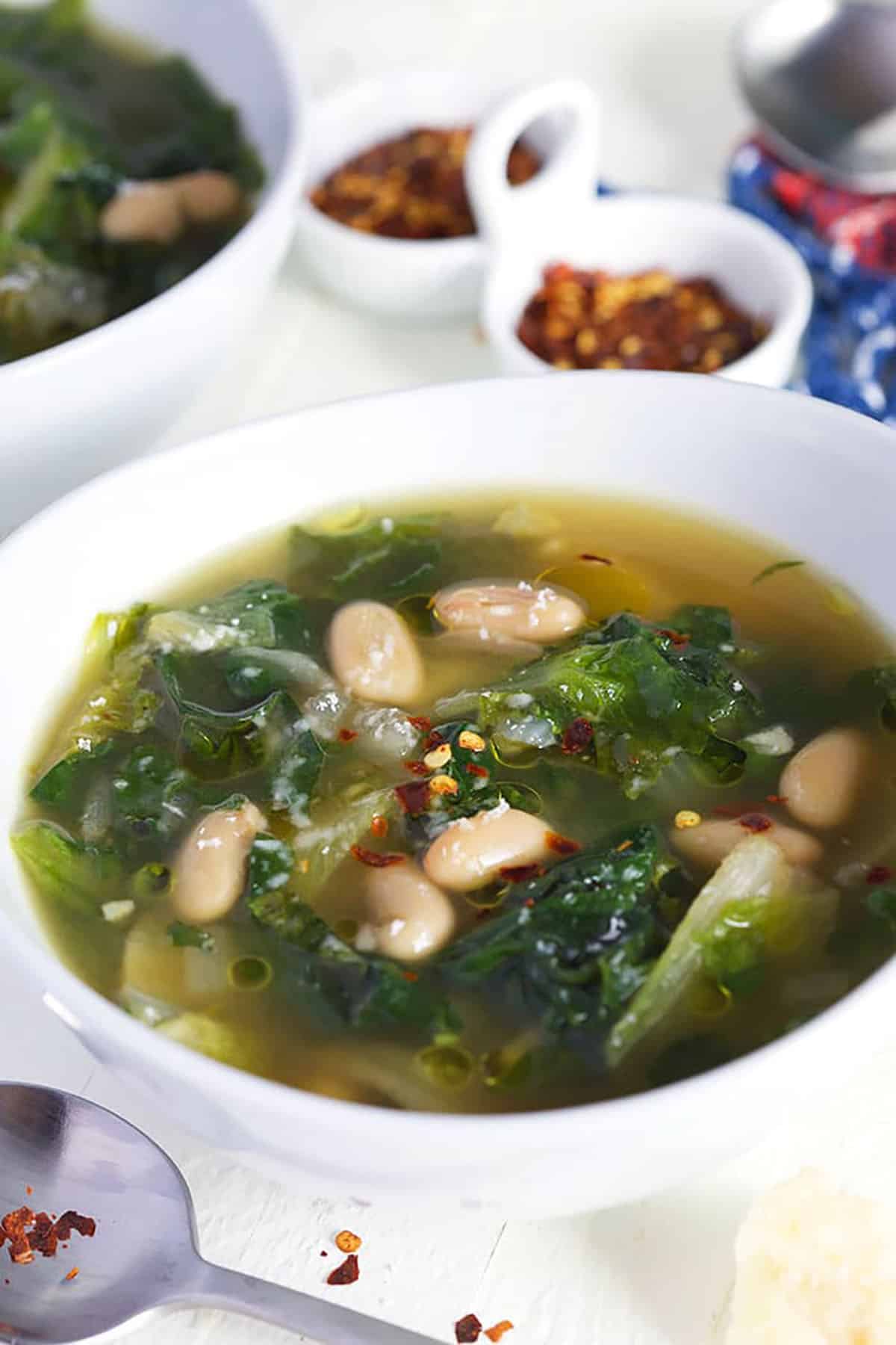 Escarole soup with beans in a white bowl with crushed red peppers in a white dish.