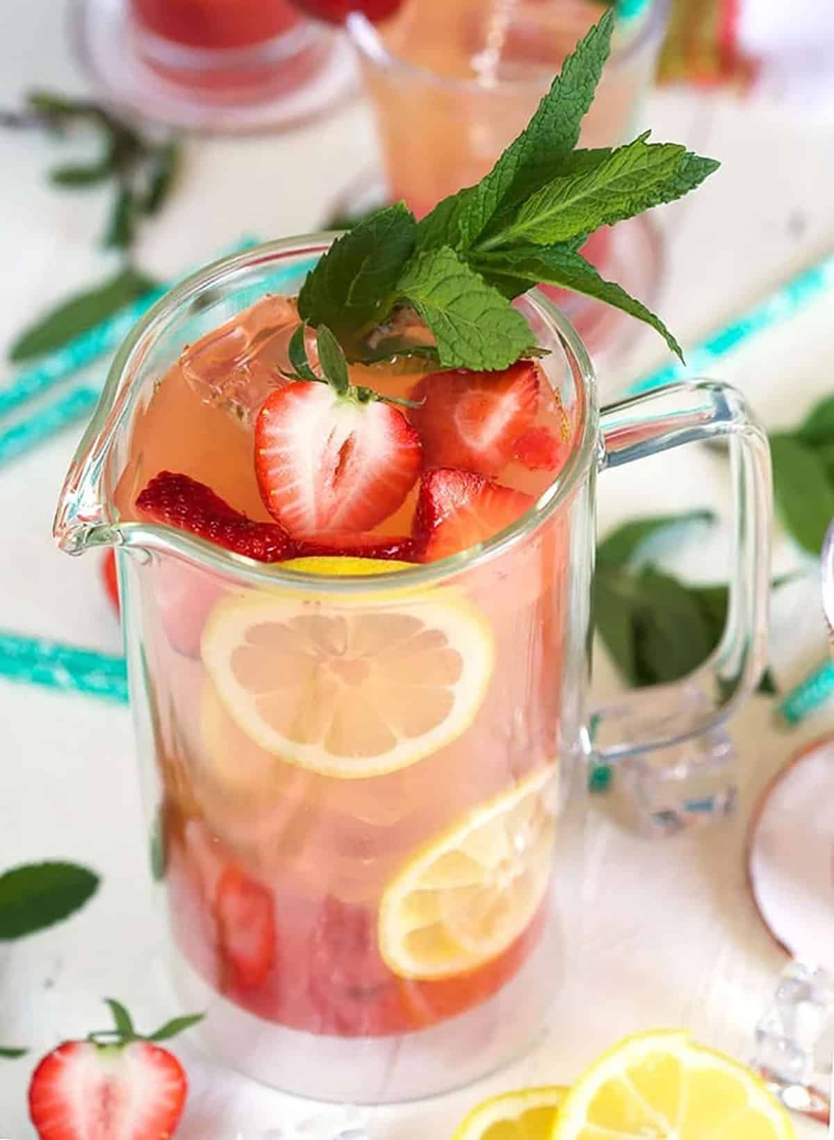 strawberry lemonade in a clear pitcher with strawberries and lemon as a garnish.