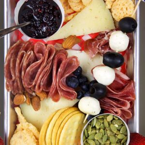 Overhead shot of charcuterie items on a stainless steel board with a red and white paper under the cheese.