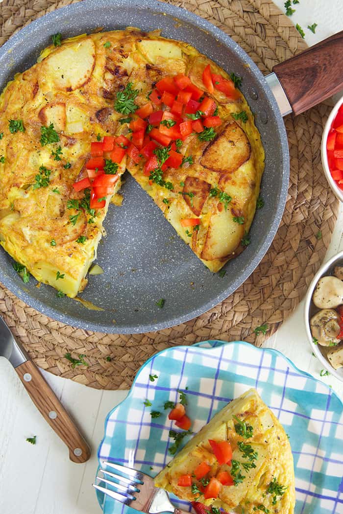 Overhead shot of Spanish omelette in a silver skillet with a wooden handle. and a slice of omelette on a plaid plate.