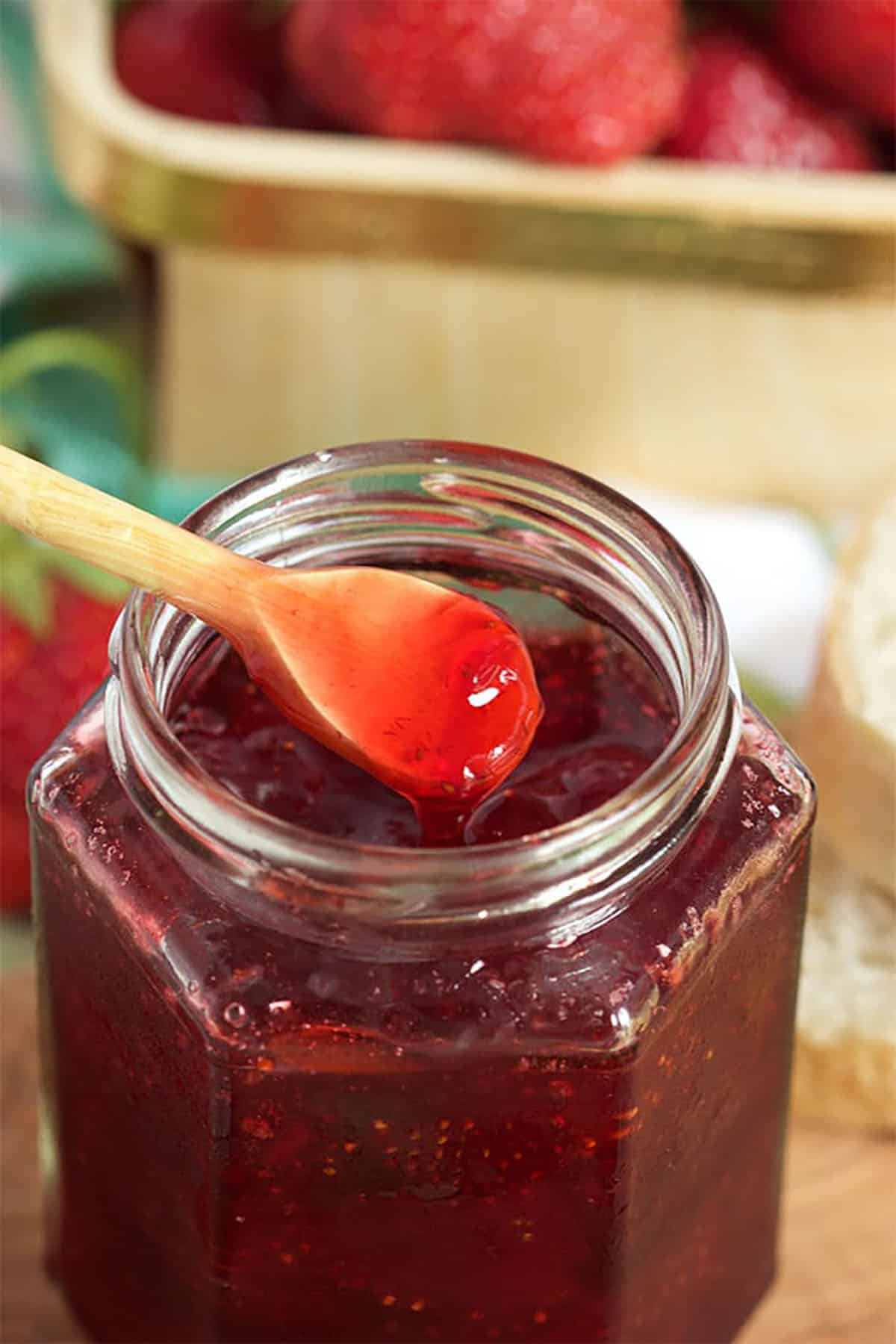 Strawberry jam in a jelly jar with a wooden spoon dipped into it.