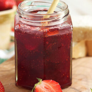 Strawberry jam in a jelly jar with a wooden spoon and two slices strawberries in front on a wooden board.