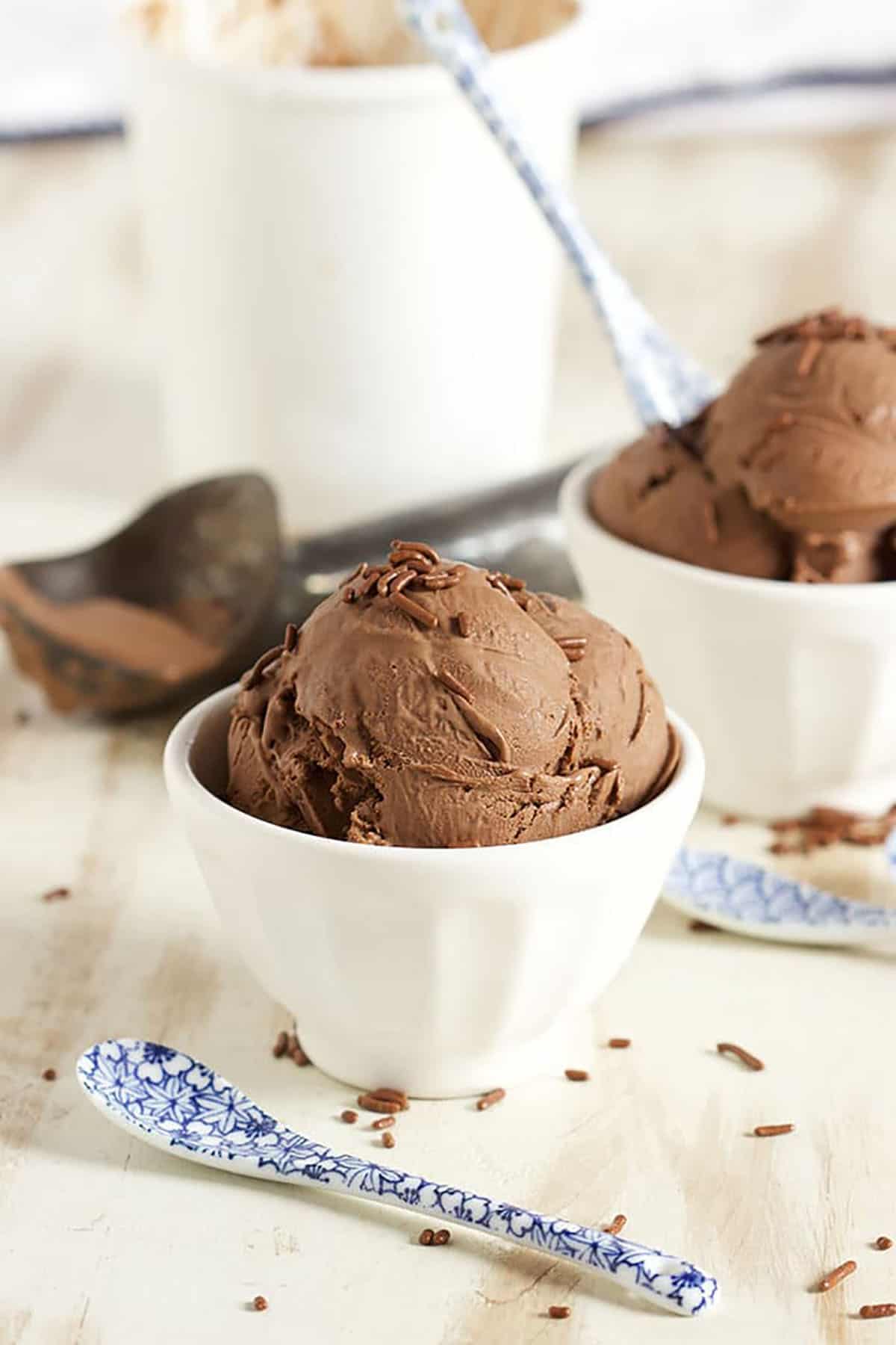 Chocolate ice cream in a white bowl on a white washed board with a blue and white spoon in front of it.