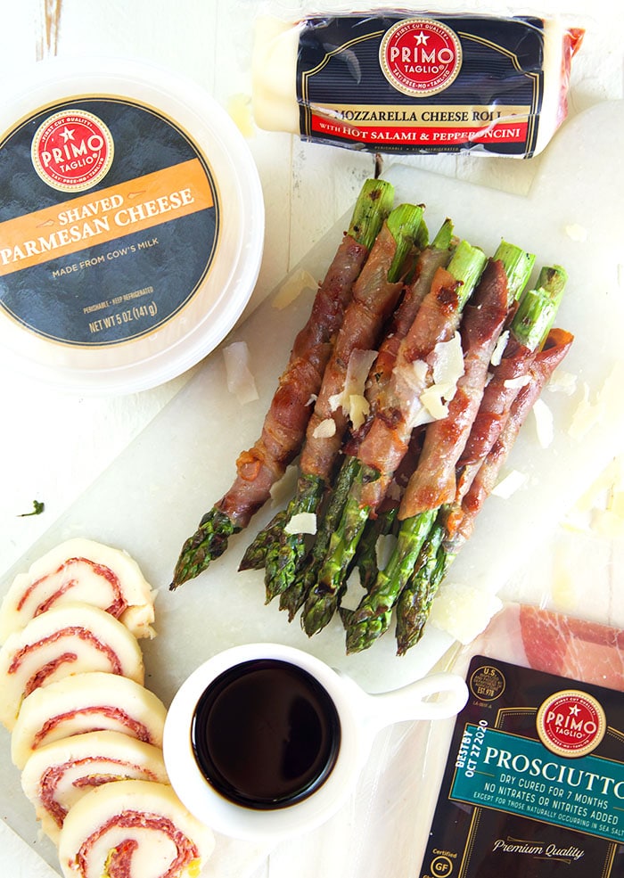 Overhead shot of prosciutto wrapped asparagus with primo taglio products on a white board.