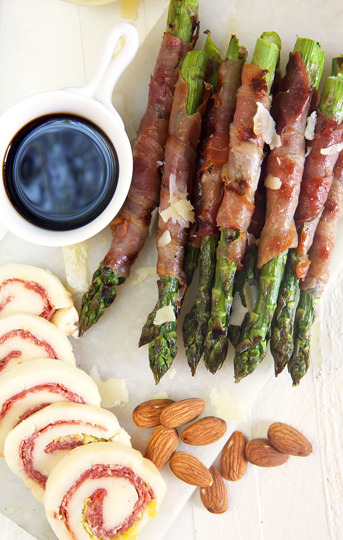 Overhead shot of prosciutto wrapped asparagus on a white board with sliced mozzarella pinwheels and almonds.