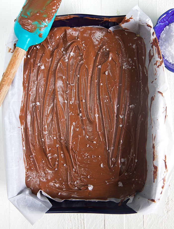 Overhead shot of scotcheroos in a baking dish with chocolate being spread with a blue spatula.