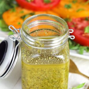 Basil Pesto Vinaigrette in a glass jar with a plate of tomatoes in the background.
