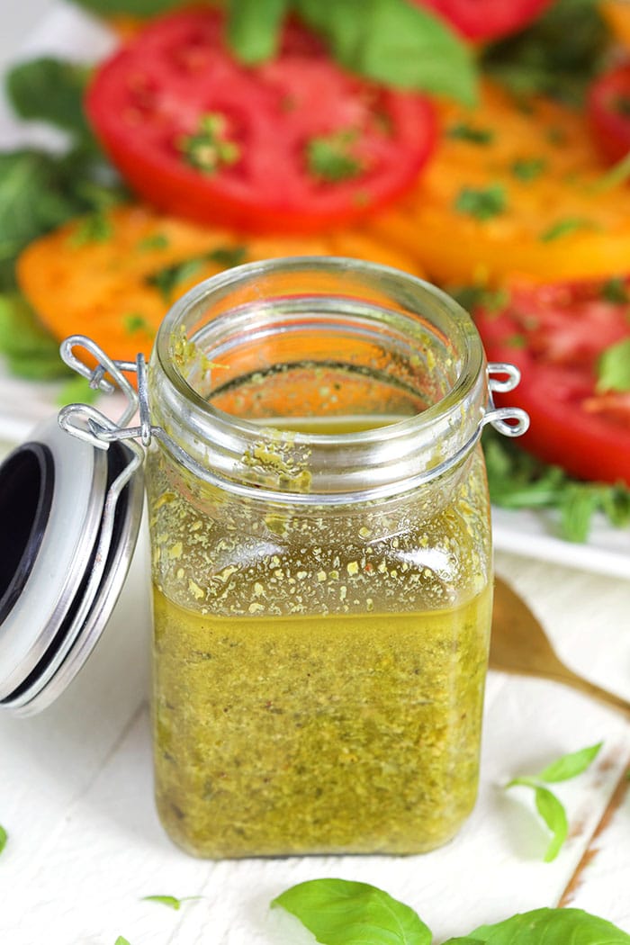 Basil Pesto Vinaigrette in a glass jar with a plate of tomatoes in the background.