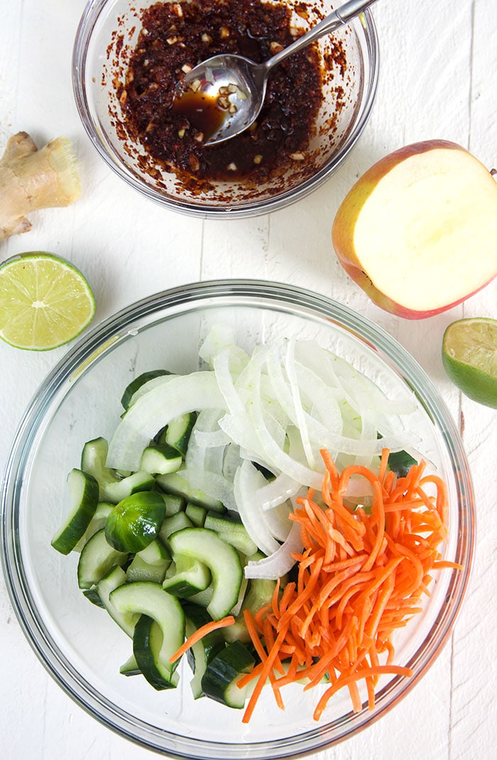 Overhead shot of cucumbers carrots and onion in a bowl with a bowl of kimchi sauce.