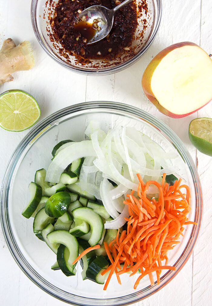 Overhead shot of cucumbers carrots and onion in a bowl with a bowl of kimchi sauce.