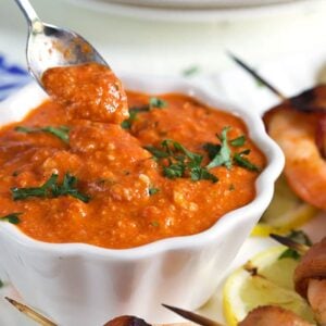 Romesco Sauce in a white bowl with a silver spoon and grilled shrimp on the side.