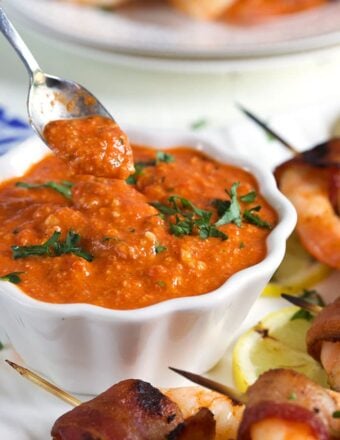 Romesco Sauce in a white bowl with a silver spoon and grilled shrimp on the side.