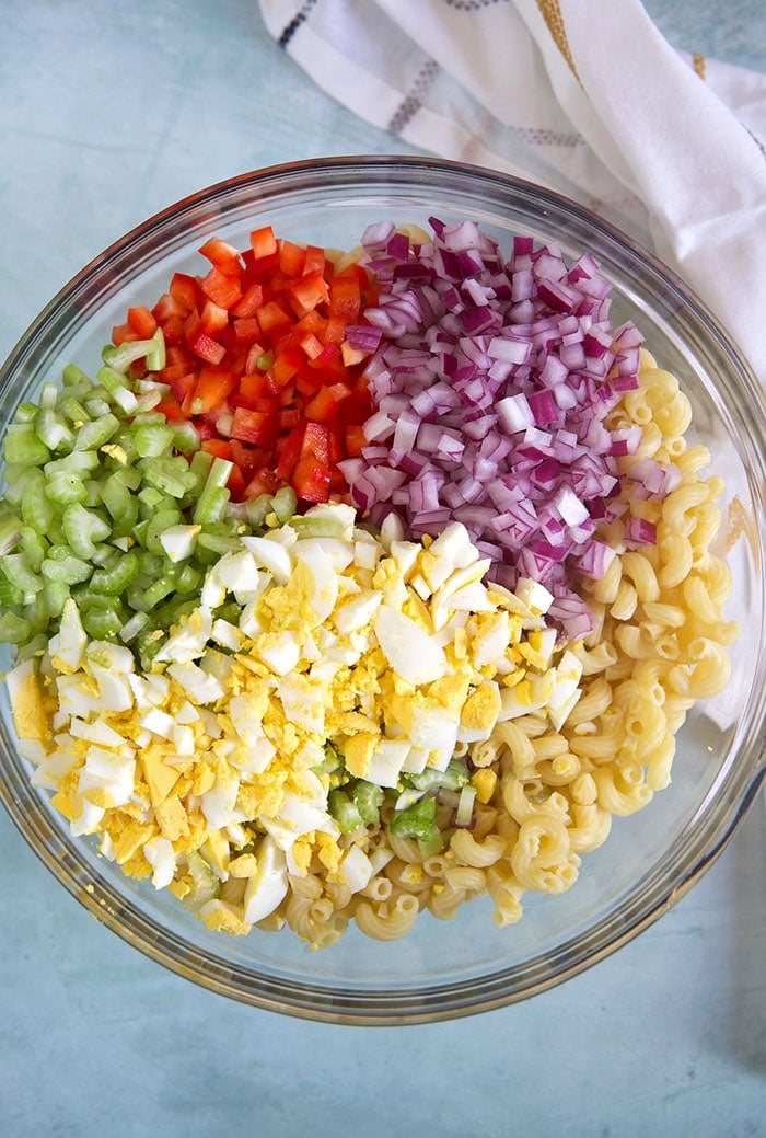 Overhead shot of ingredients for macaroni salad in a glass bowl on a blue background.