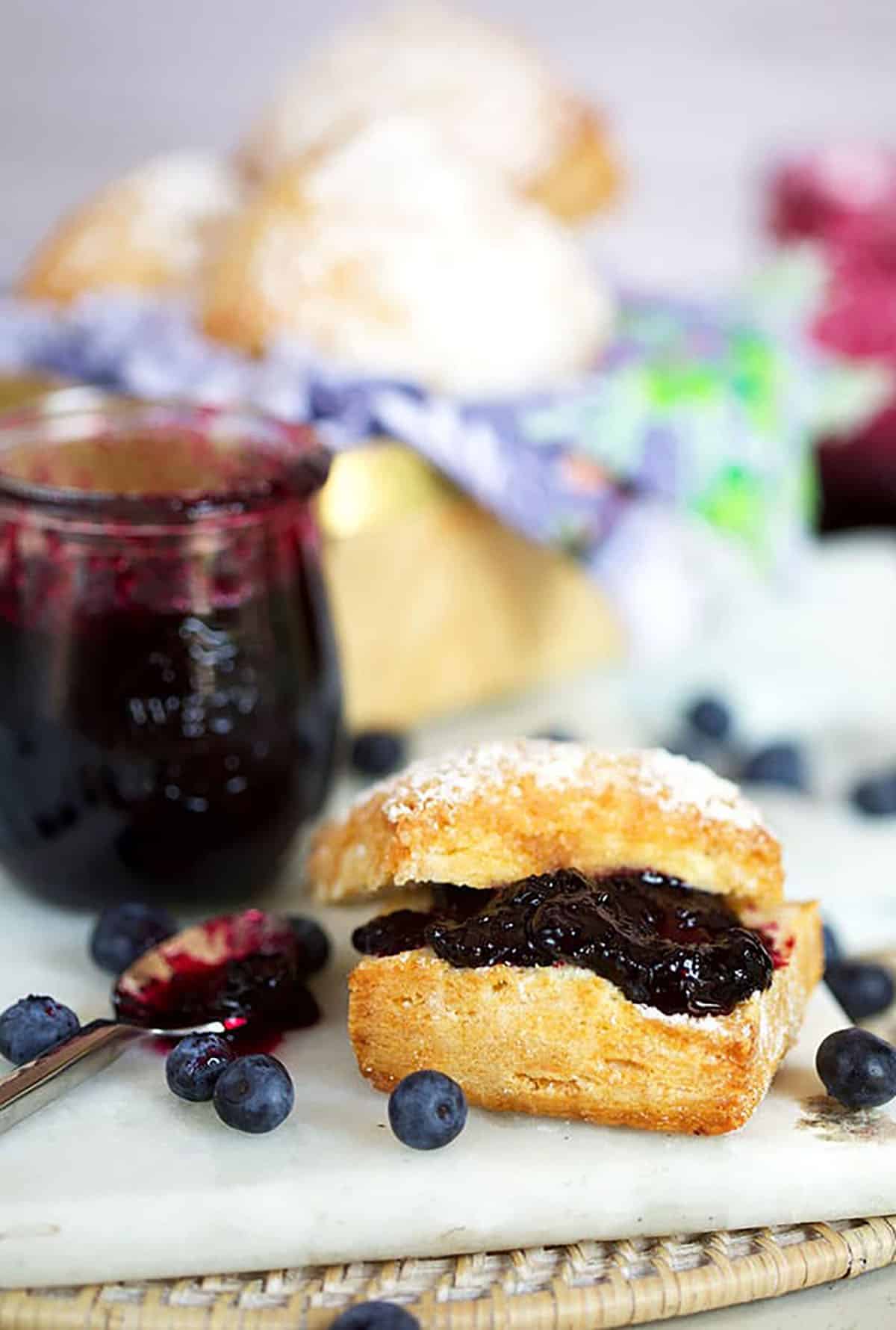 Blueberry jam on a biscuit with a jar of blueberry jam in the background on a white marble board
