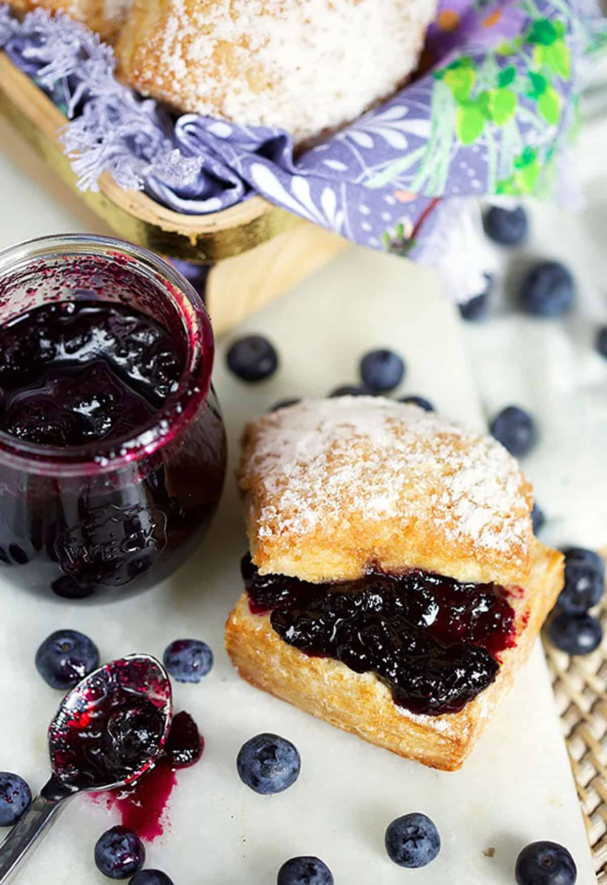 Close up shot of blueberry jam on a biscuit with a jar of jam and a messy spoon on a white board 