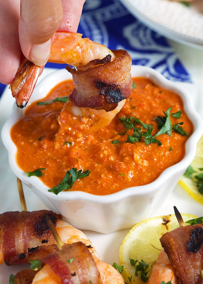 Bacon wrapped shrimp being dipped into a bowl of romesco sauce in a white ceramic bowl with a lemon slice next to it on a white background with a blue and white napkin in the background.