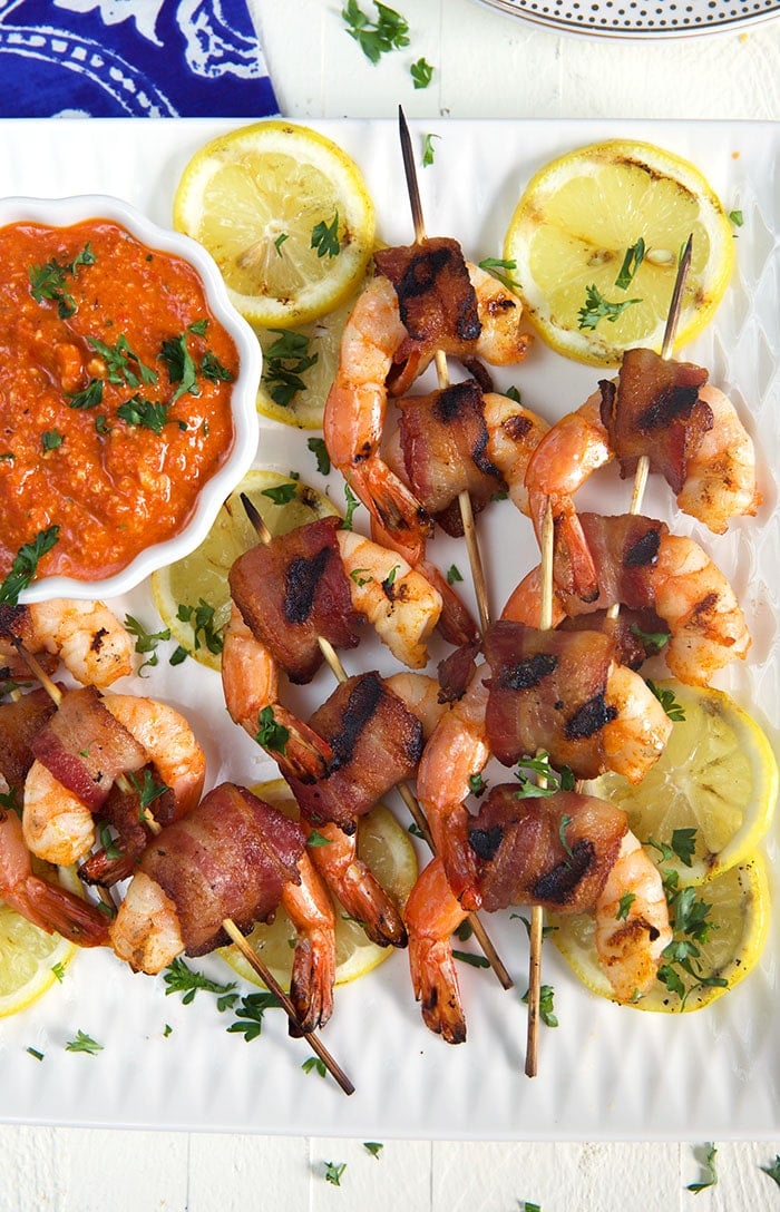 Overhead shot of bacon wrapped shrimp on bamboo skewers on a white platter with red romesco sauce in a white bowl.
