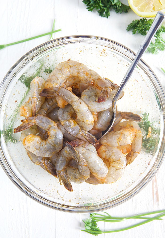 Overhead shot of shrimp in a glass bowl with a spoon on a white background and parsley sprigs on the side.