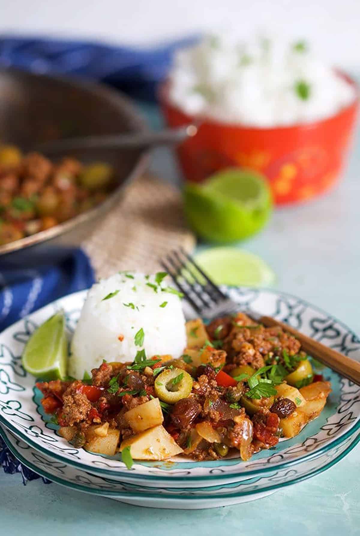 Picadillo on a plate with rice and a wood spoon and a bowl of rice in the backgound.