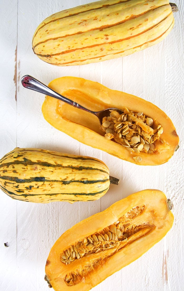 Overhead shot of delicata squash cut in half with a spoon scooping out the seeds on a white background.