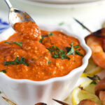 Romesco Sauce in a white bowl with a silver spoon and grilled shrimp on the side.