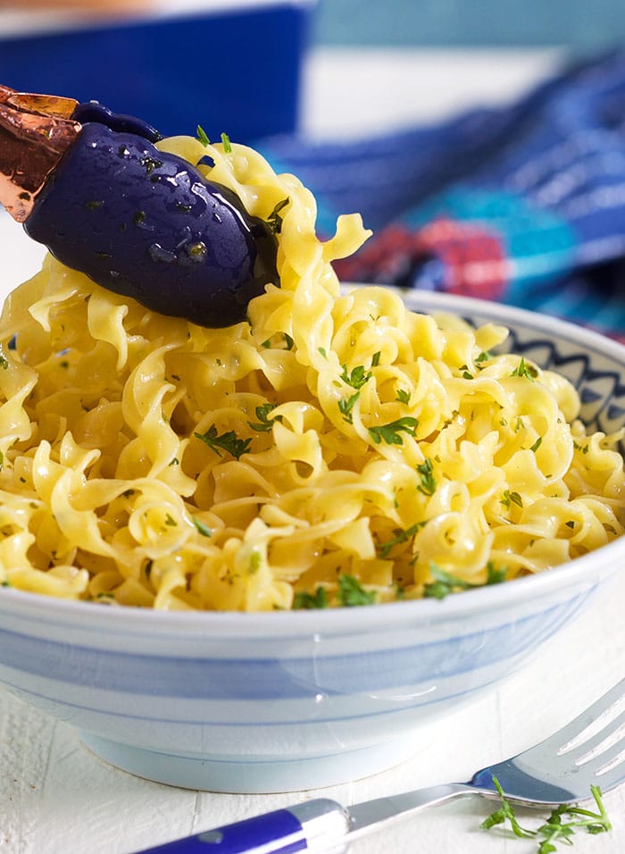 Close up of buttered noodles being served with blue tongs.