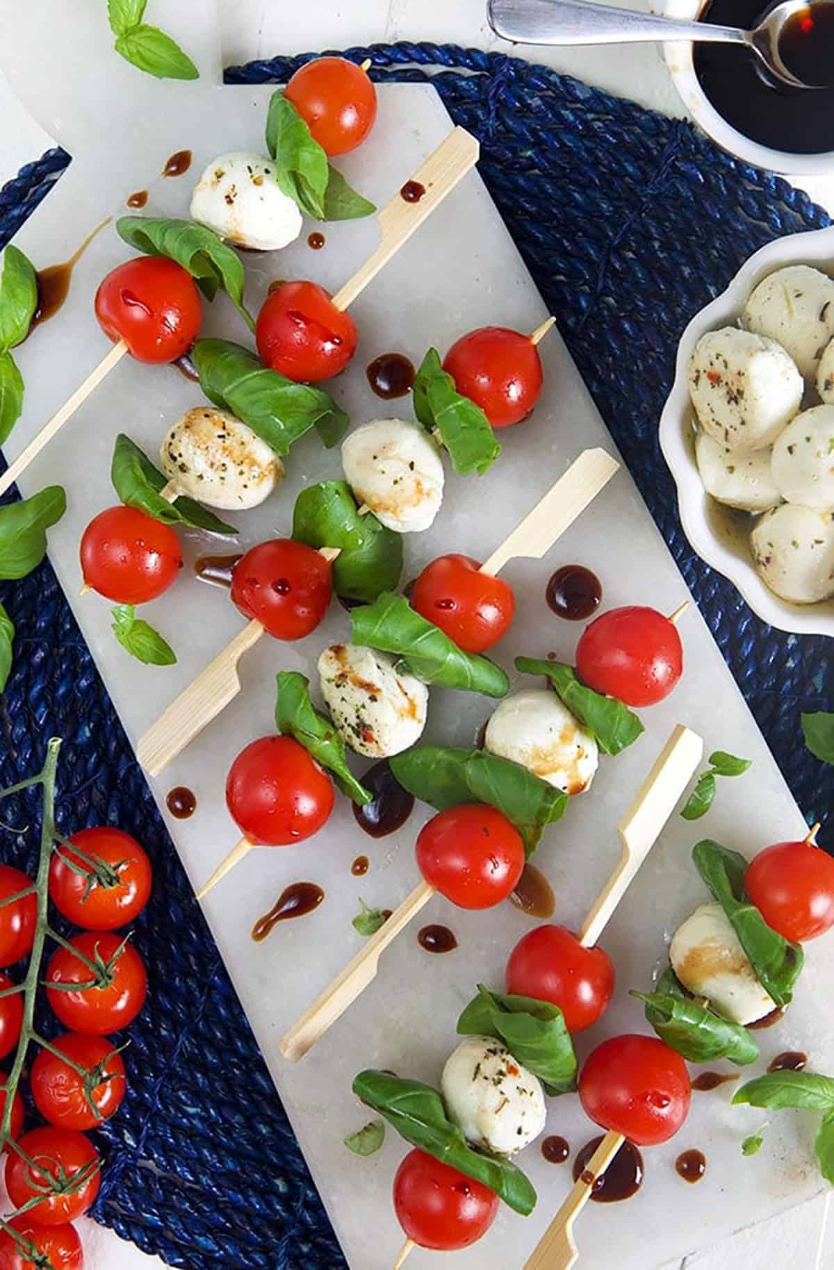 Caprese Salad Skewers on a marble tray with mozzarella balls in a white bowl.