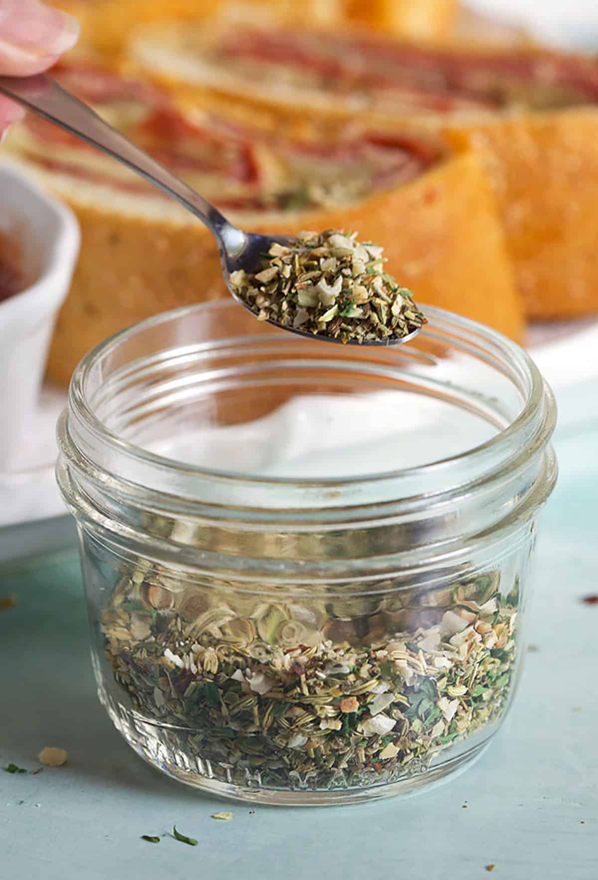 Jar with Pizza Seasoning and a spoon with a scoop of seasoning on it.