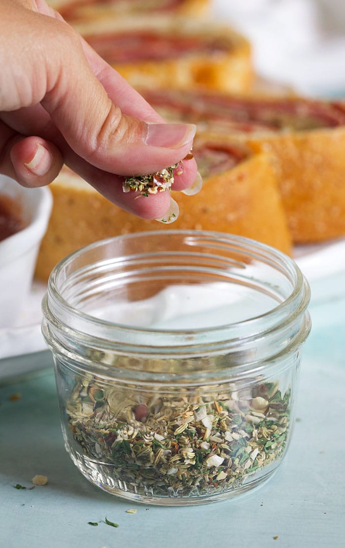 Pizza seasoning in a little jar with a hand grabbing a pinch of seasoning.