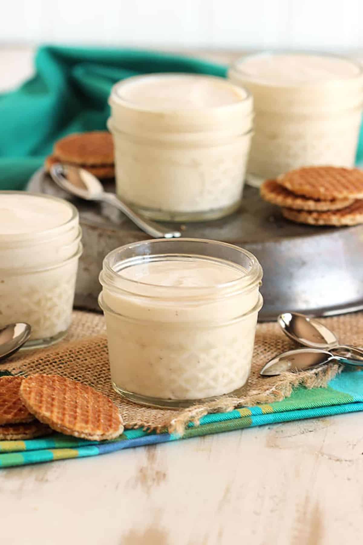 Four small jars with vanilla pudding in them and mini stroopwafels on the side.
