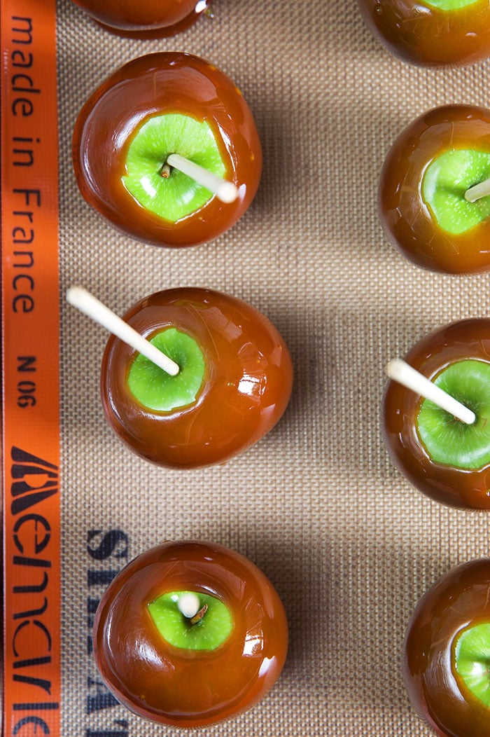 6 caramel apples on a baking sheet on a silicon baking mat.
