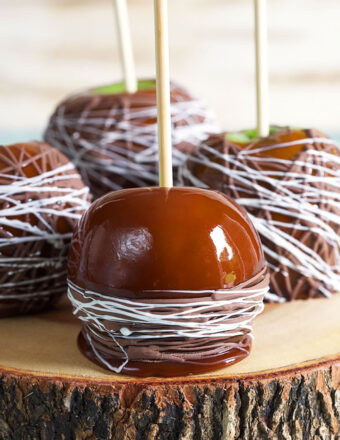 Four caramel apples drizzled with chocolate on a slab of wood.