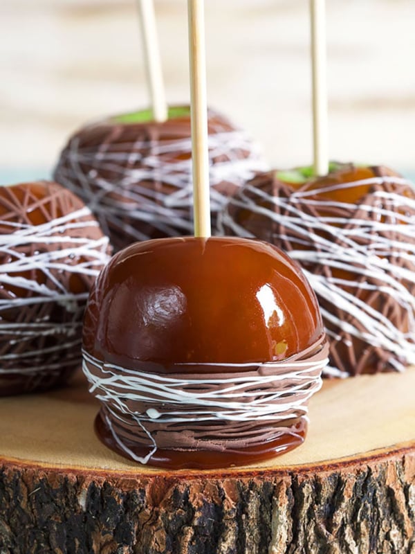 Four caramel apples drizzled with chocolate on a slab of wood.