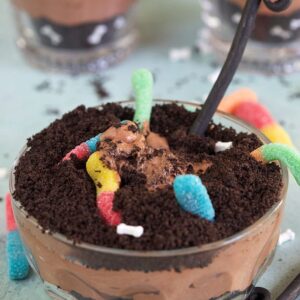 Dirt cake with gummy worms in a glass bowl with a spoon on a blue background.