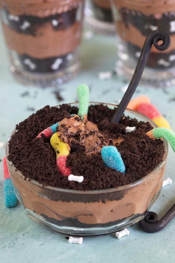 Dirt cake with gummy worms in a glass bowl with a spoon on a blue background.